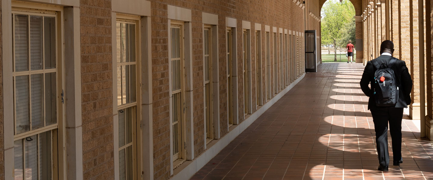 Man in a suit and backpack walks through an external corridor on a college campus.